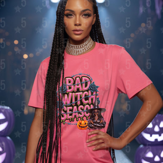 Woman wearing a pink 'Bad Witch Season' t-shirt with Halloween-themed graphics against a festive background.