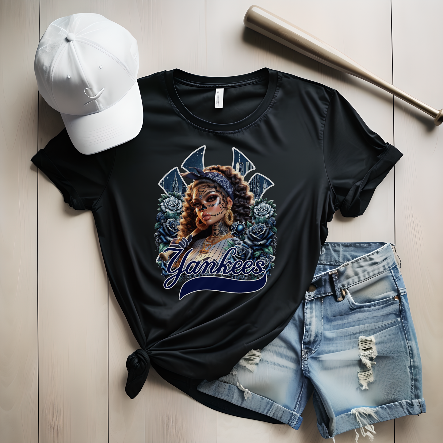 Black t-shirt with dia de los muertos 'Yankees' design, white cap, and denim shorts on a light wooden surface.