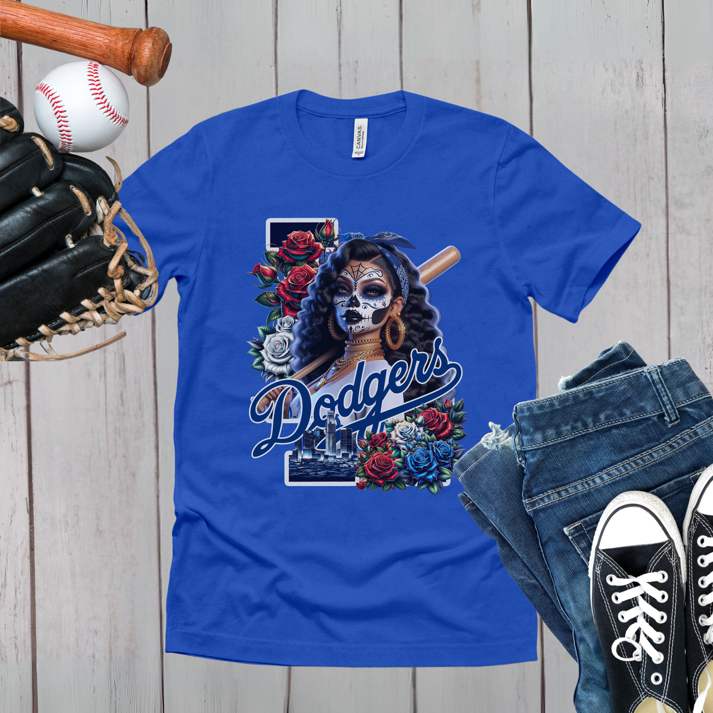 Blue t-shirt with 'Dodgers' dia de los muertos design on a wooden surface with baseball and jeans.