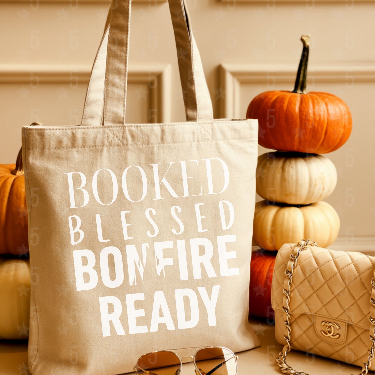 Beige tote bag with text 'BOOKED BLESSED BONFIRE READY' in front of pumpkins and a quilted handbag.