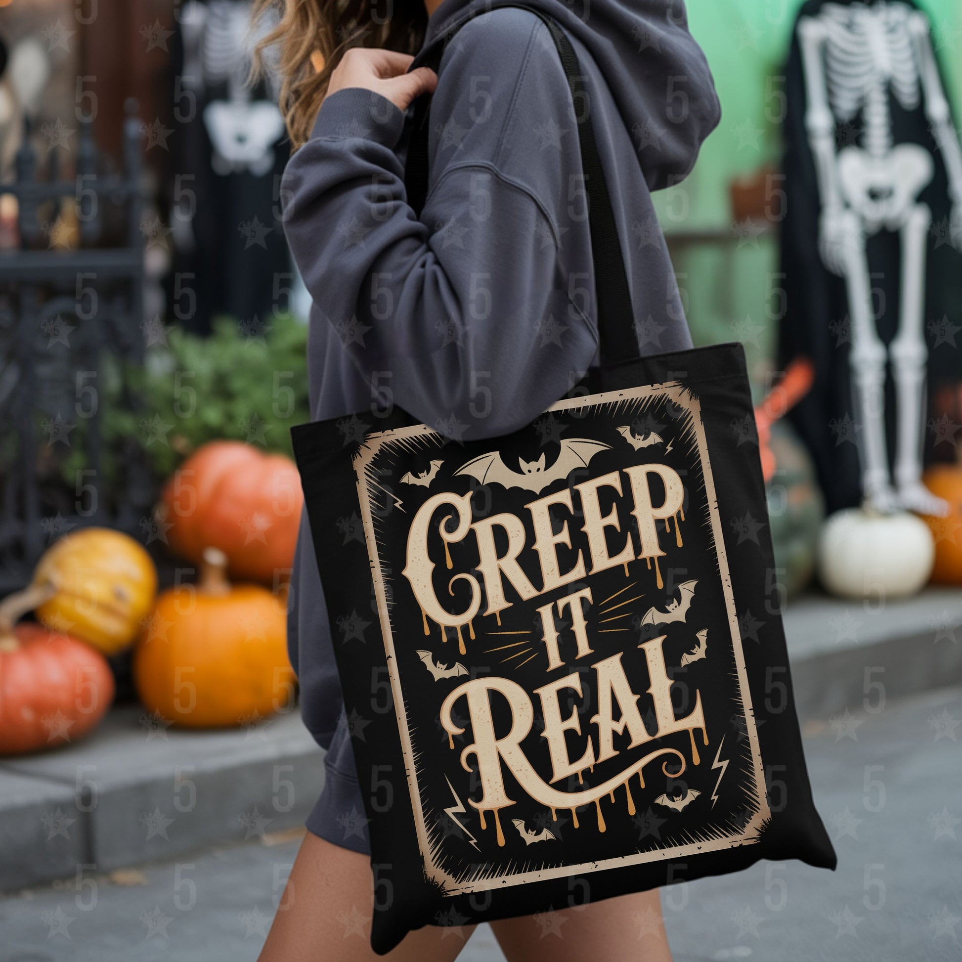 Person holding a tote bag with 'Creep It Real' text in a Halloween-themed setting with pumpkins and skeletons.