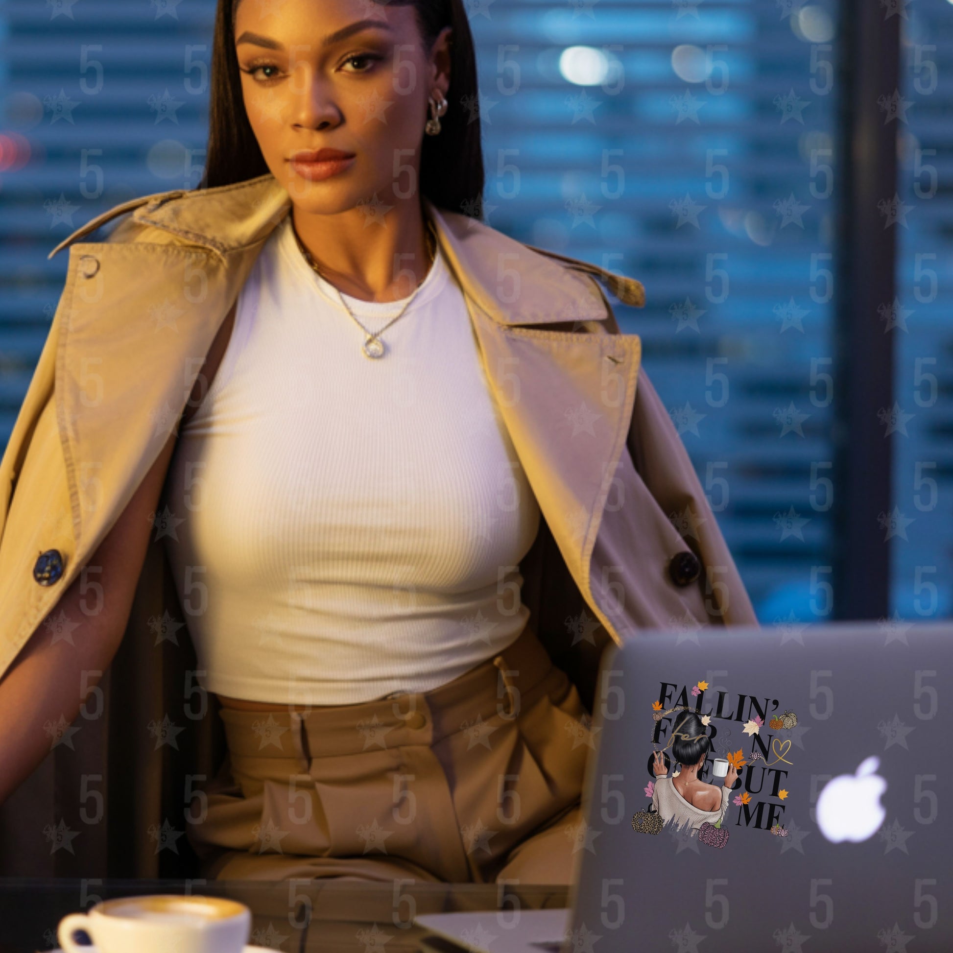 Woman in a beige trench coat sitting at a desk with a laptop and coffee cup, with a blurred background.