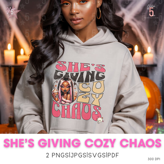 Person wearing a hoodie with 'SHE'S GIVING COZY CHAOS' text and graphics, with candles and pumpkins in the background.
