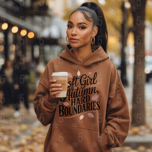 Woman wearing a brown hoodie with text, holding a coffee cup in an autumn setting.