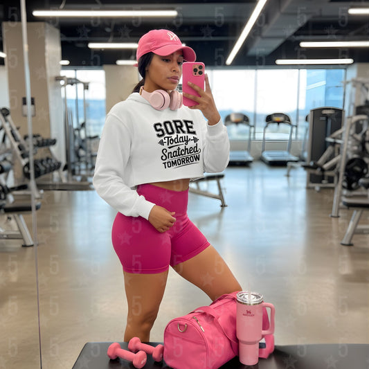 Person in a gym wearing a white cropped hoodie and pink shorts, taking a mirror selfie.