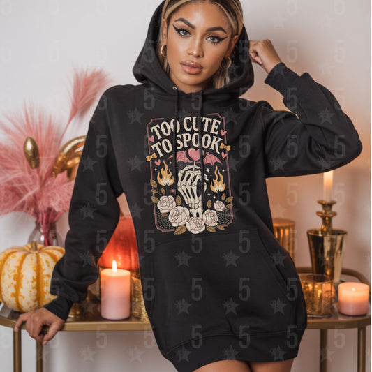 Woman wearing a black hoodie with 'Too Cute To Spook' design in a decorative setting.
