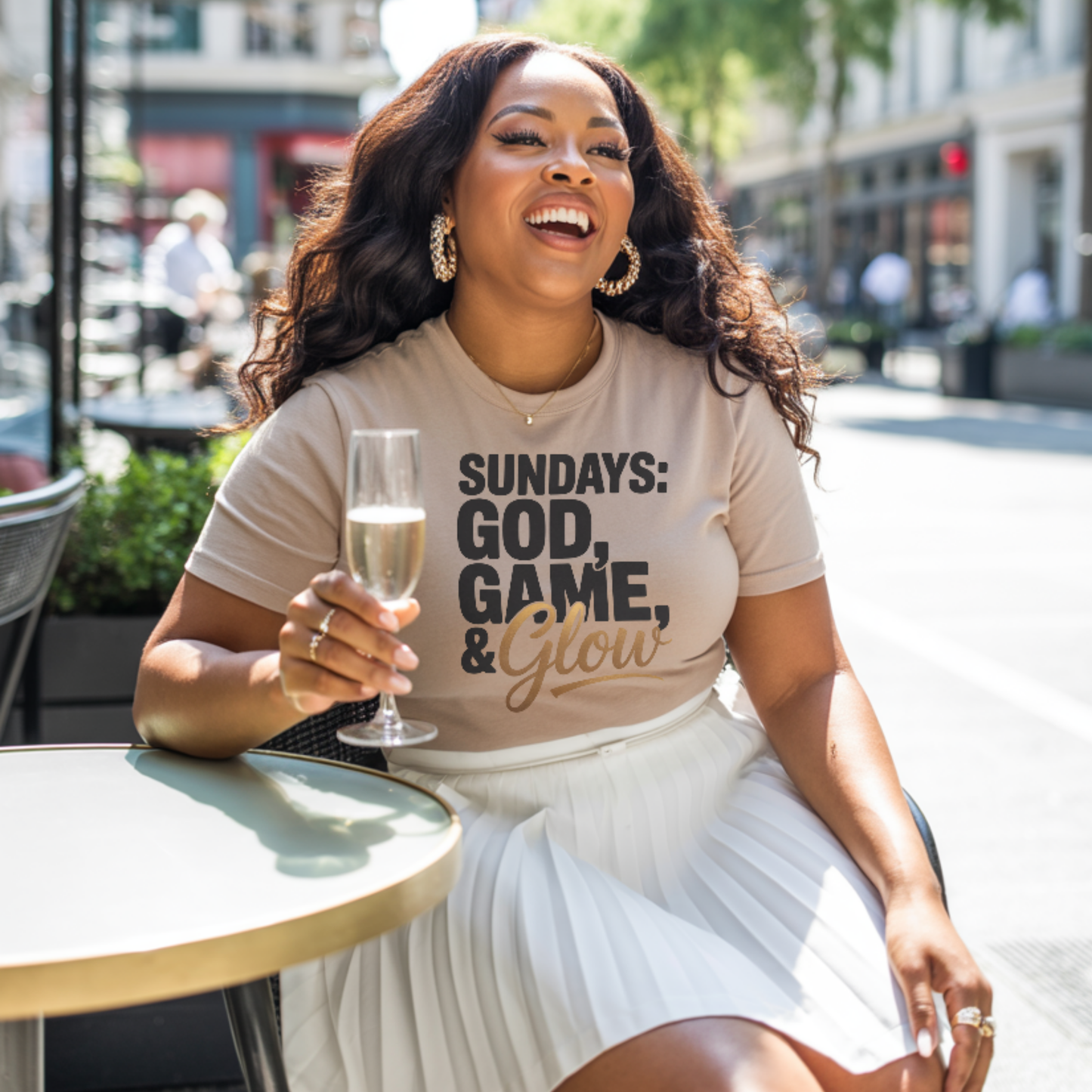 Woman sitting outdoors holding a glass of champagne, wearing a t-shirt with sundays god game glow design.