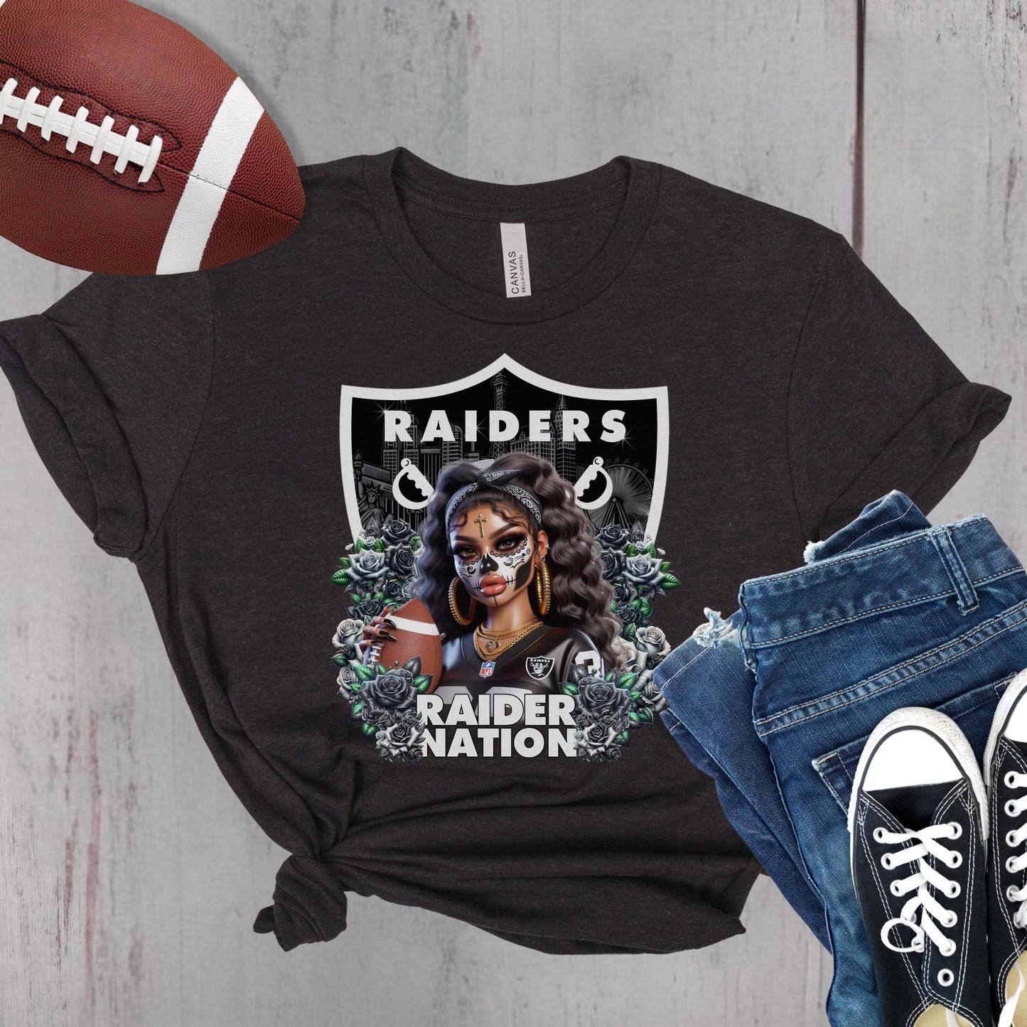 Black t-shirt with a dia de los muertos Raiders design, football, jeans, and sneakers on a wooden surface.