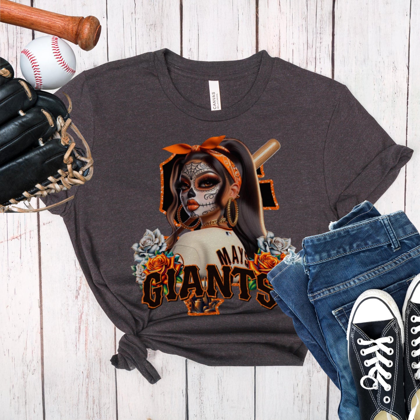 Dark gray t-shirt with a sf giants dia de los muertos design of a woman in a mask and hat, surrounded by roses, on a wooden surface with a baseball, bat, glove, jeans, and black sneakers.