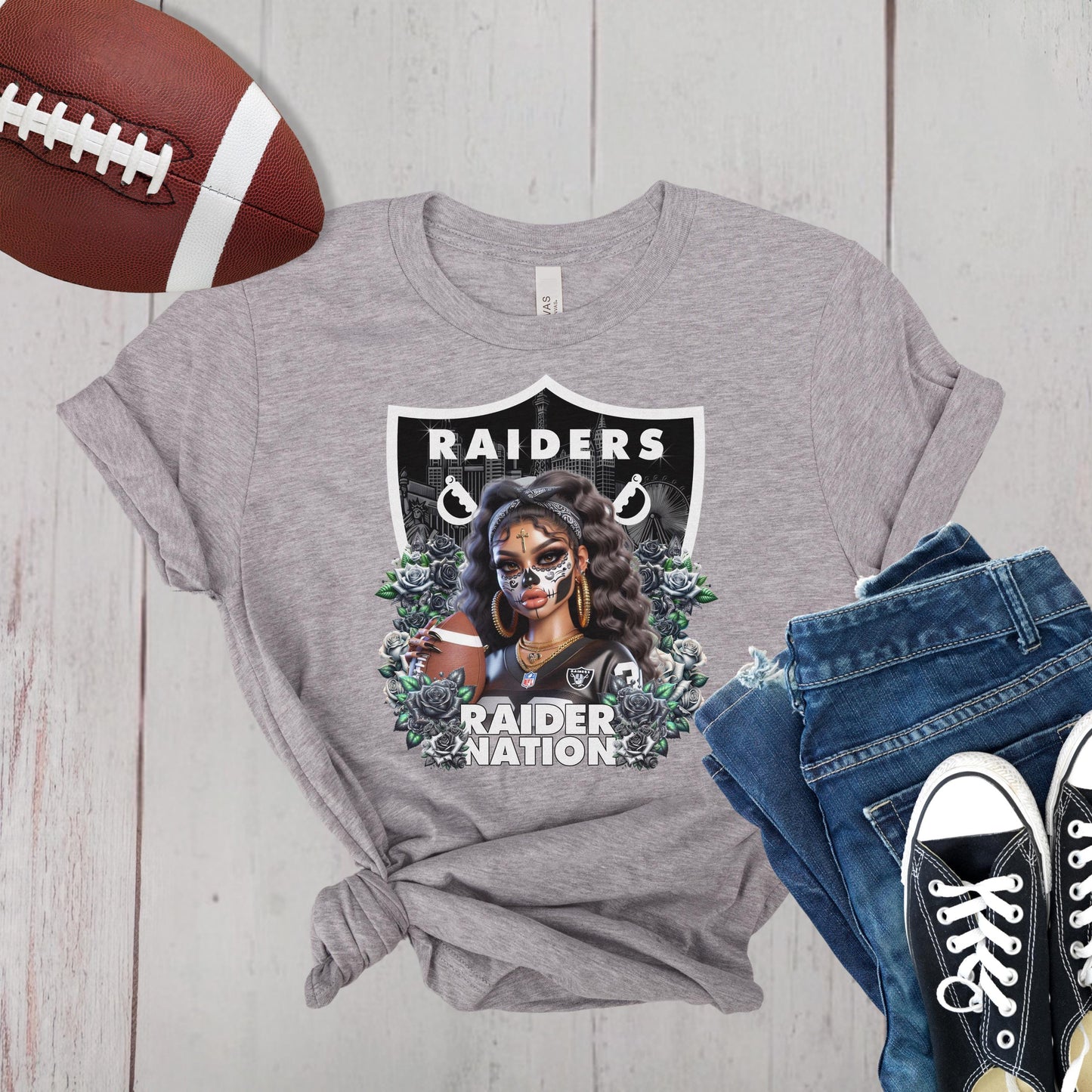 Gray t-shirt with a dia de los muertos Raiders design, football, jeans, and sneakers on a wooden surface