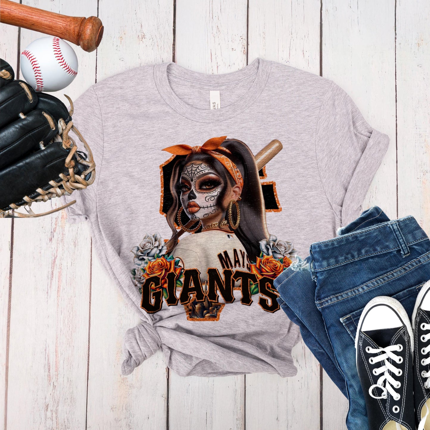 Gray t-shirt with a sf giants dia de los muertos design of a woman with a baseball bat, surrounded by sports equipment on a wooden surface.