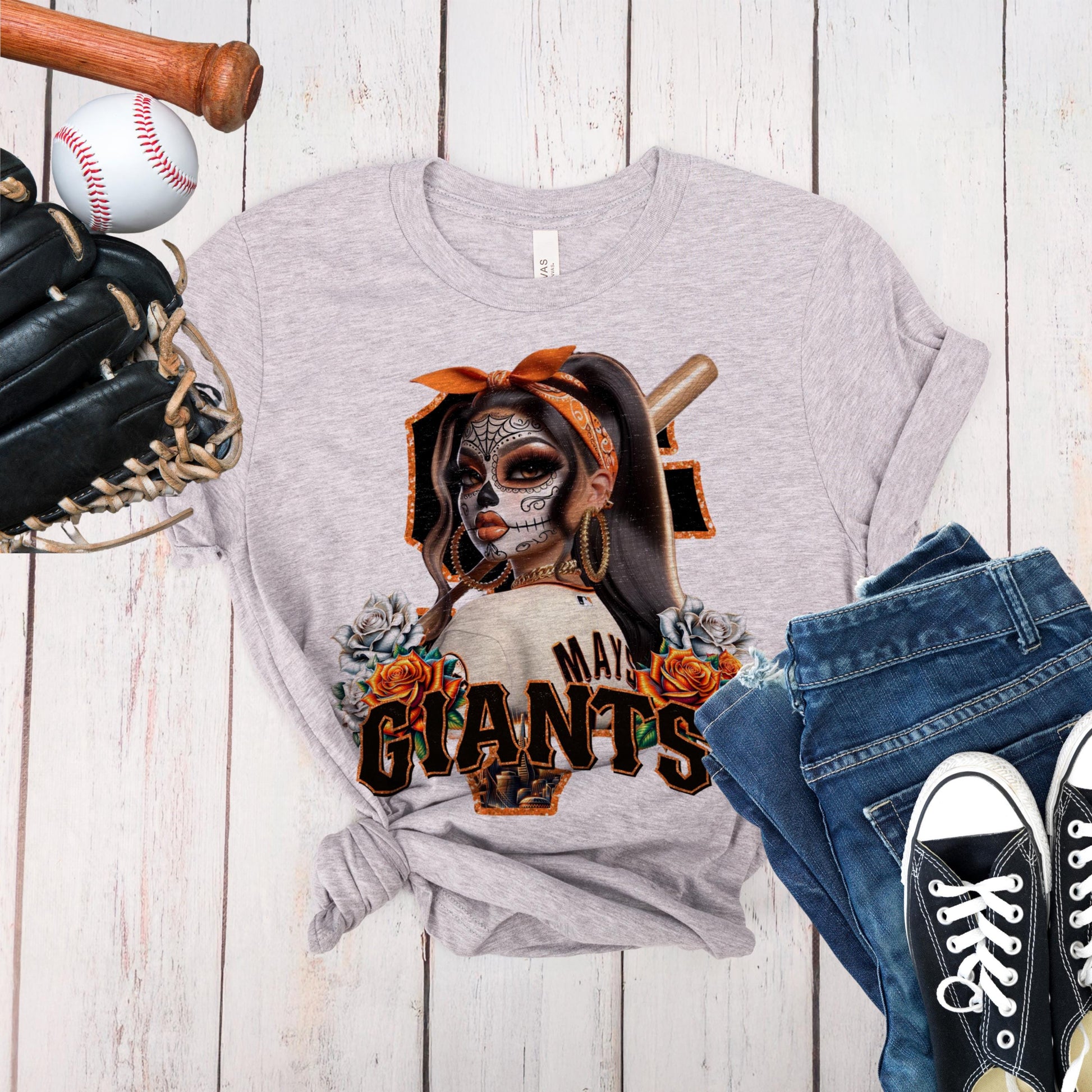 Gray t-shirt with a sf giants dia de los muertos design of a woman with a baseball bat, surrounded by sports equipment on a wooden surface.