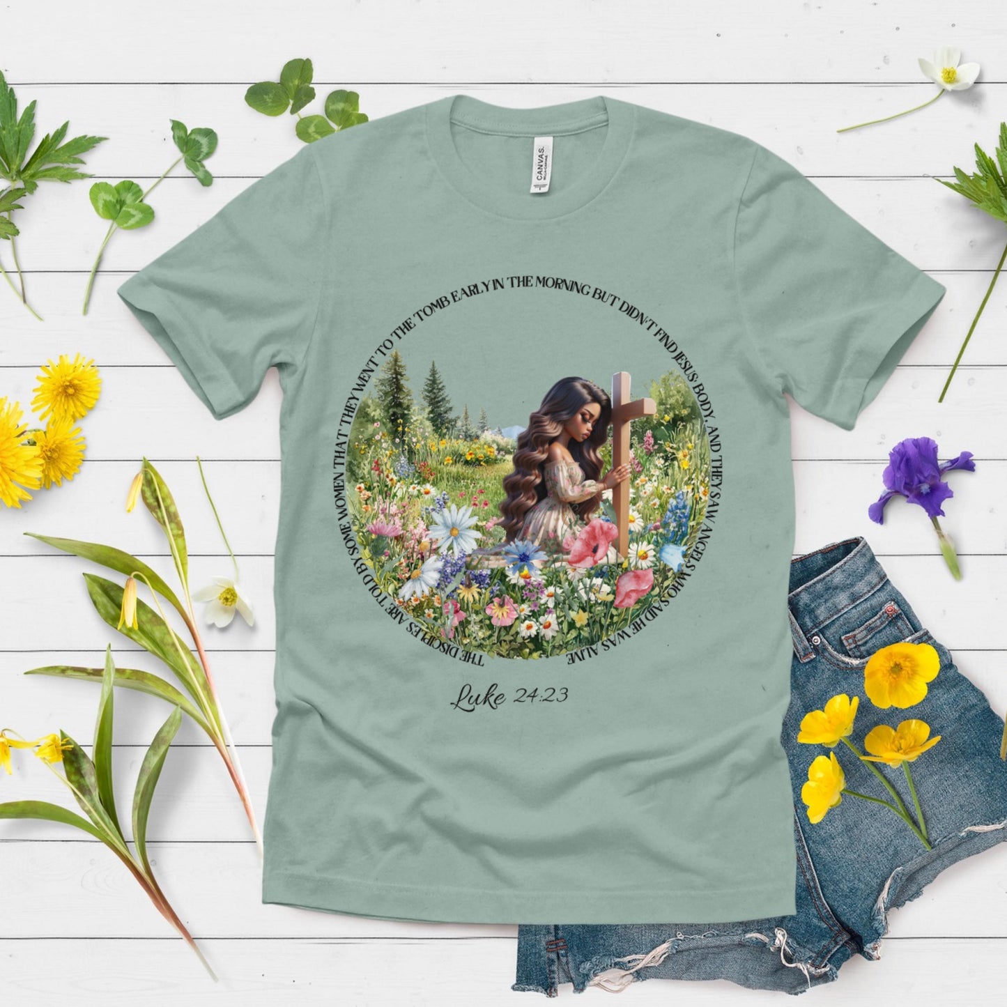 Green t-shirt with a printed design of a woman with flowers and a cross, surrounded by flowers on a wooden surface.