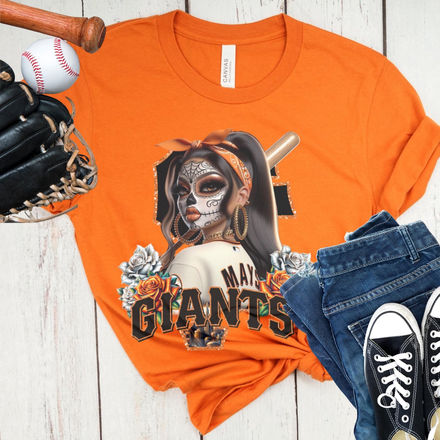 Orange t-shirt with a sf giants dia de los muertos design of a woman with a skull makeup and 'Giants' text, surrounded by sports equipment on a wooden surface.