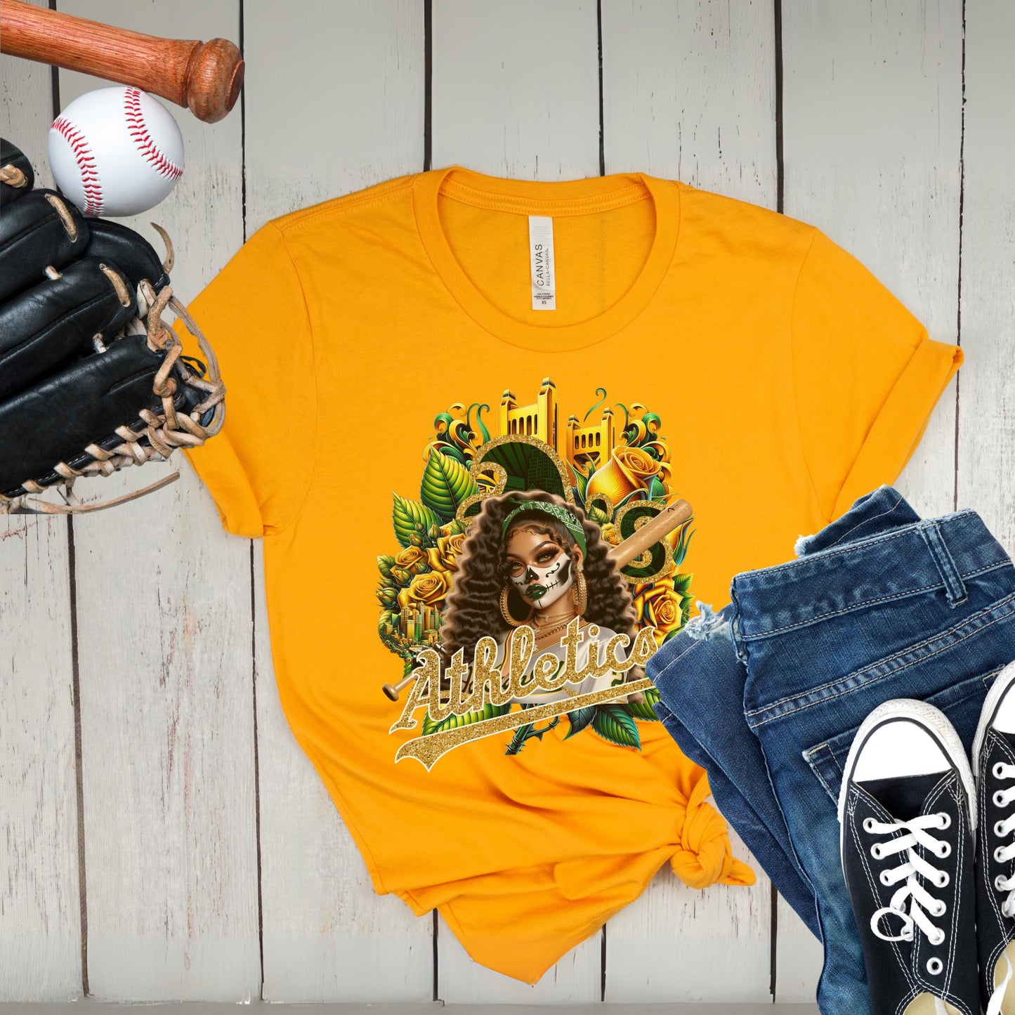 Yellow t-shirt with a graphic design of a dia de los muertos athletics, displayed with jeans and sneakers on a wooden surface.
