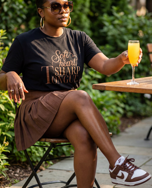 black woman drinking a mimosa in a black tee with a sassy saying "soft heart sharp tongue"