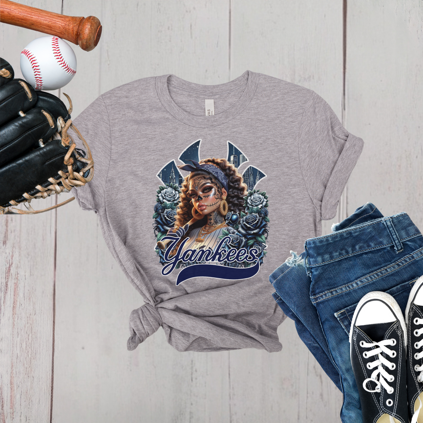 Gray t-shirt with dia de los muertos 'Yankees' design, baseball, bat, glove, and jeans on a wooden surface