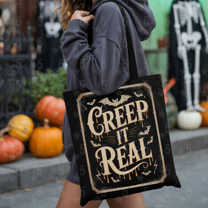 Person holding a tote bag with 'Creep It Real' text in a Halloween-themed setting with pumpkins and skeletons.