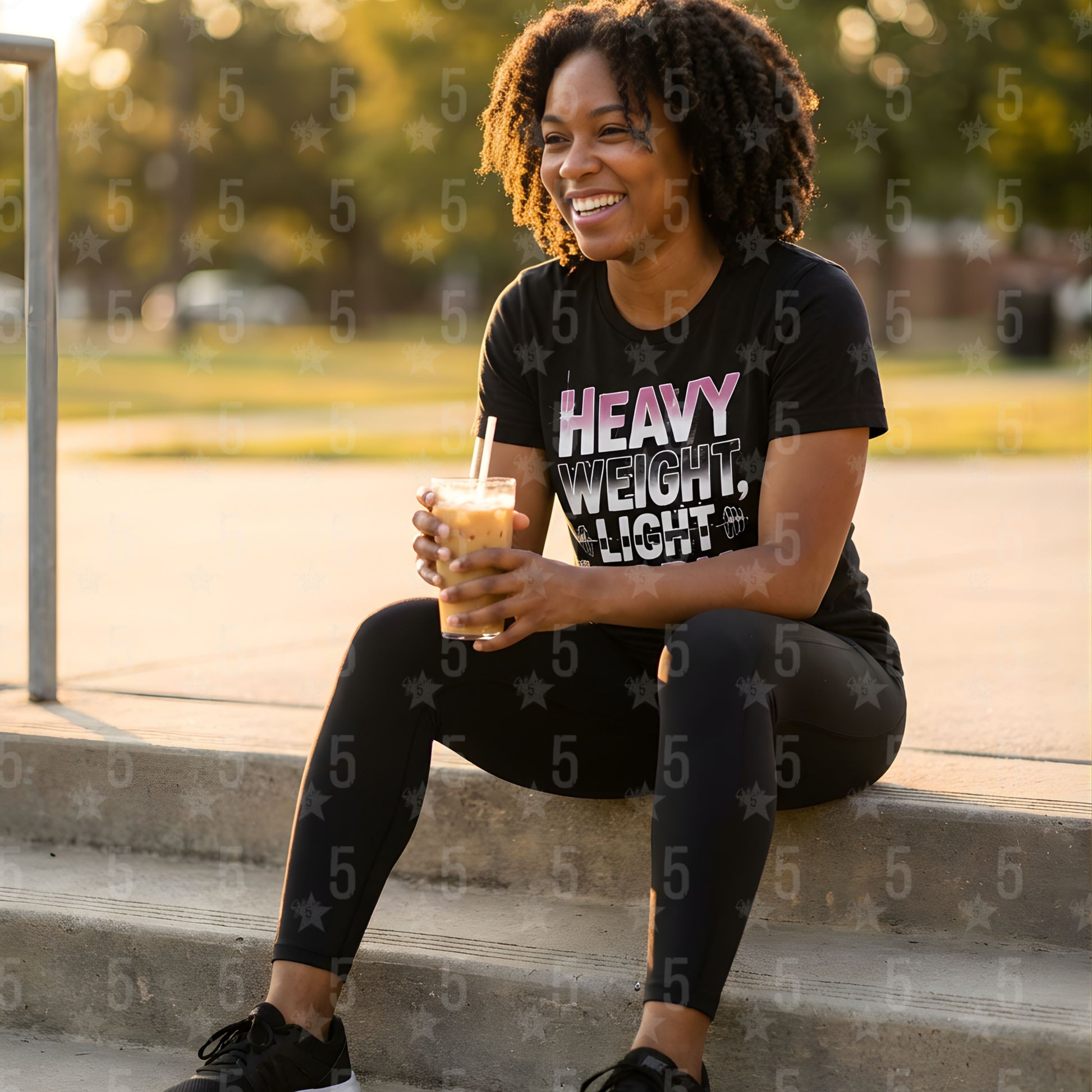 Person sitting outdoors holding a drink, wearing a black t-shirt with text and leggings.