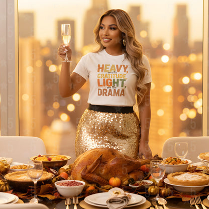 Woman in a 'Heavy is the Gratitude, Light is Drama' t-shirt standing by a table with a roasted turkey and other dishes, holding a glass of champagne.