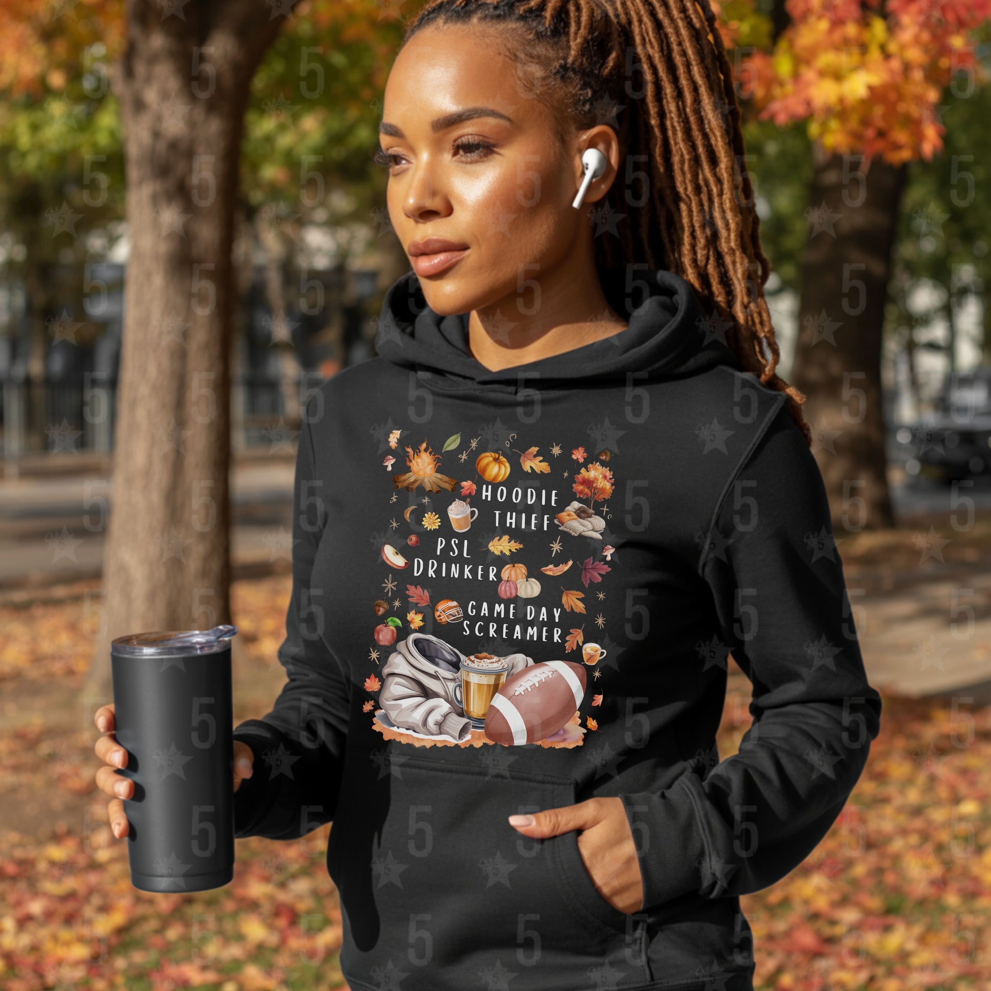 Woman wearing a black hoodie with a graphic design, holding a black tumbler in an outdoor setting with trees and fallen leaves.
