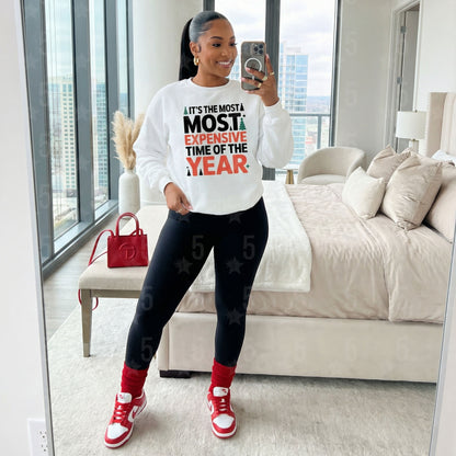 Woman taking a mirror selfie in a bedroom wearing a white sweatshirt with text, black leggings, and red sneakers.