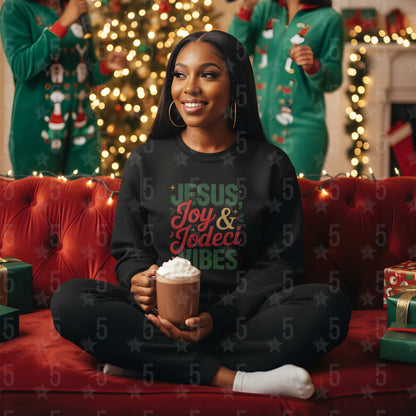 Woman wearing a black sweatshirt with Christmas-themed text, sitting on a red couch holding a mug, with festive decorations in the background.