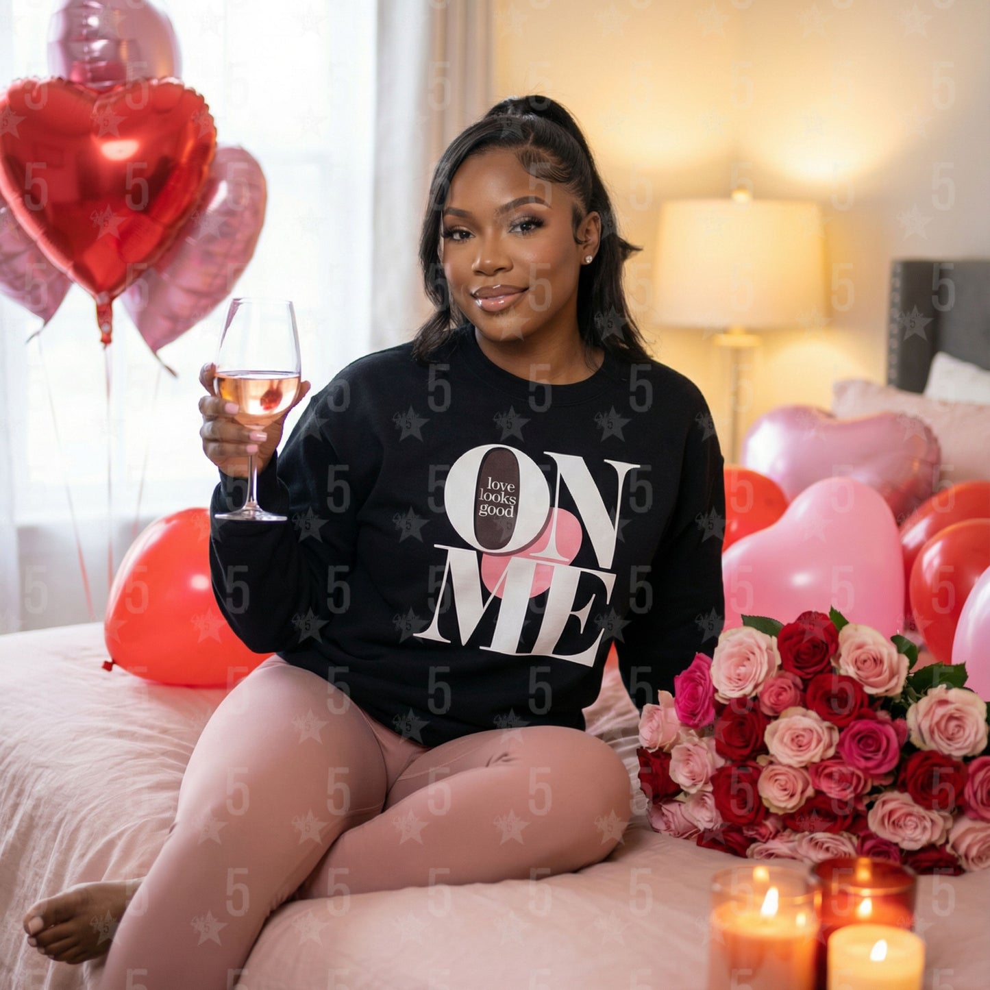 Woman sitting on a bed with balloons, flowers, and a glass of wine, wearing a black sweatshirt with text.