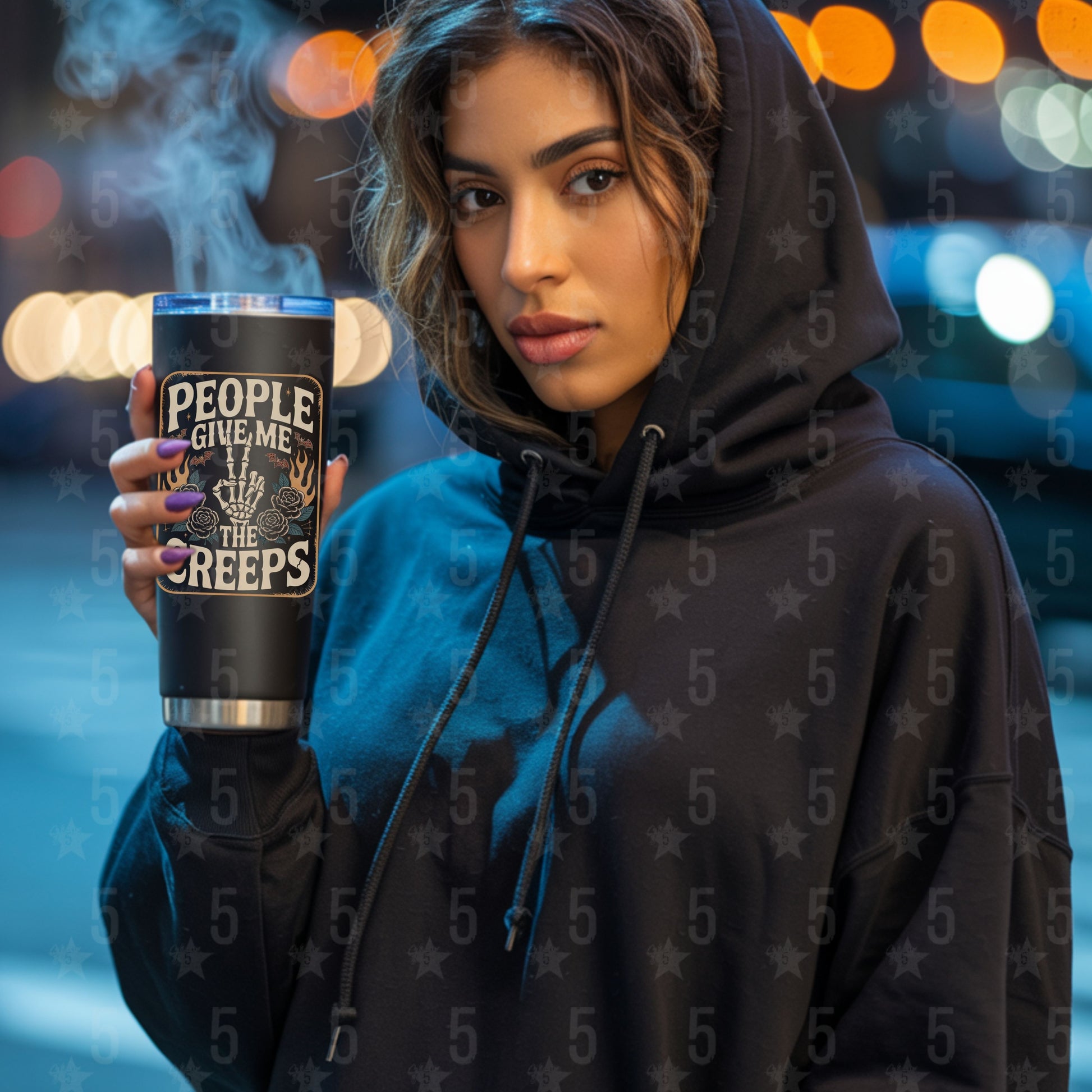Person wearing a hoodie holding a mug with text, blurred lights in the background