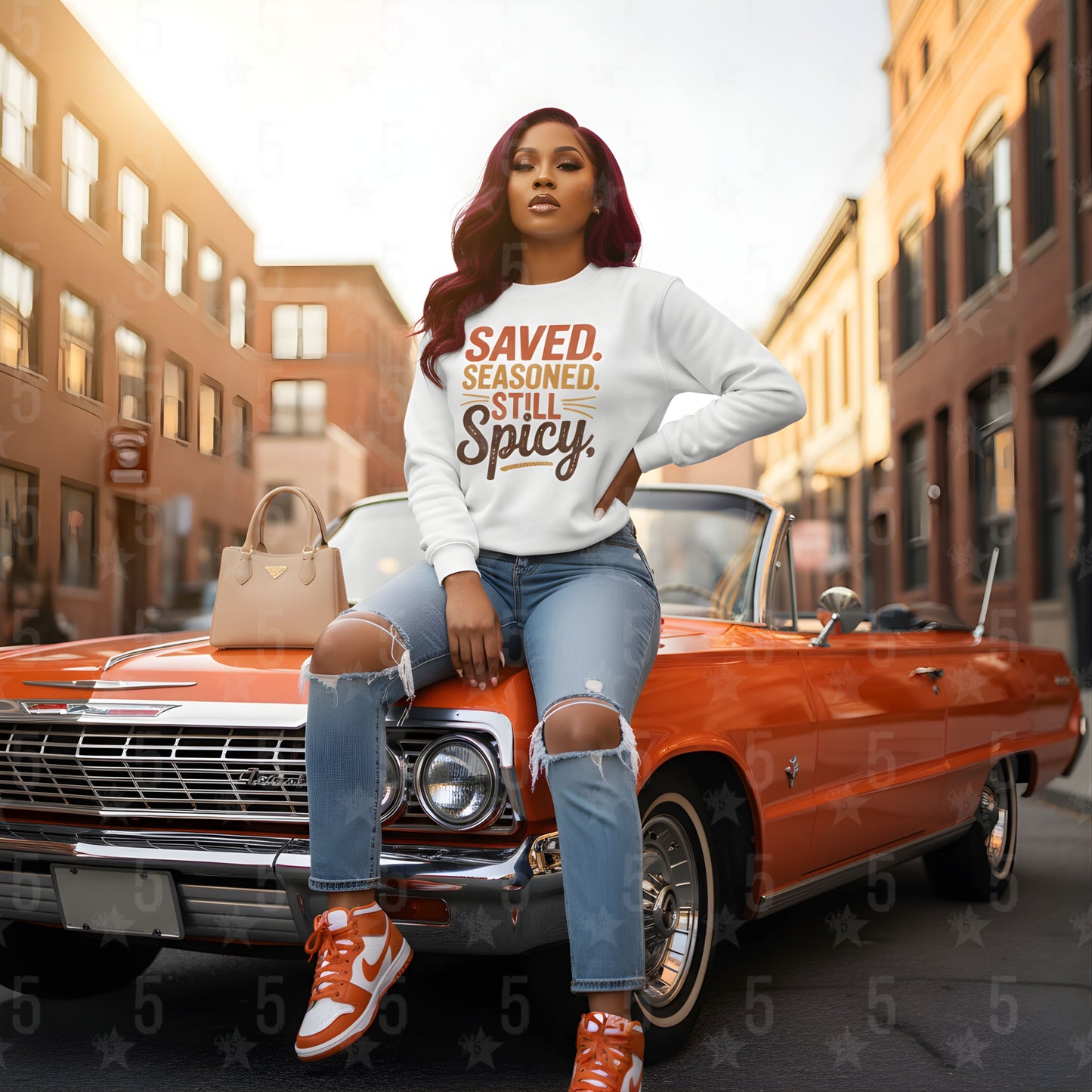 Woman sitting on a classic red car wearing a white sweatshirt with text, in an urban setting.