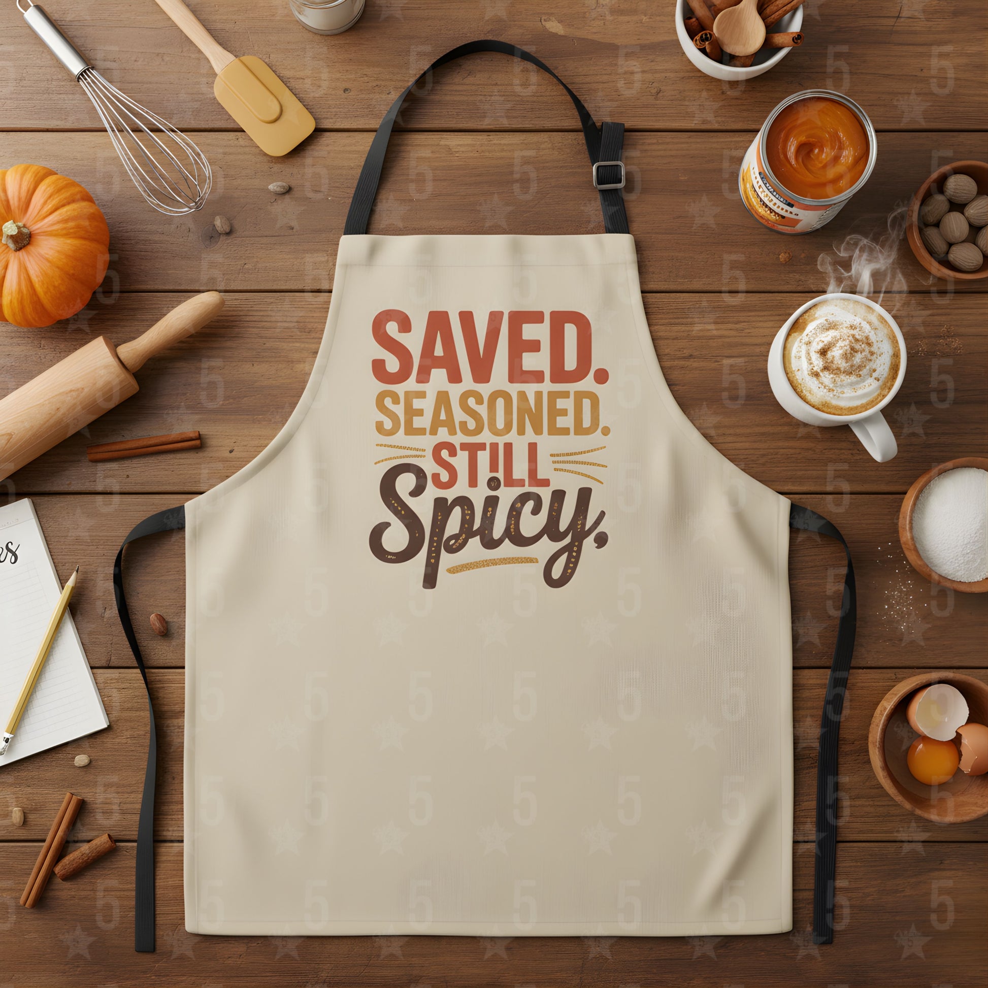 Beige apron with 'Saved. Seasoned. Still Spicy.' text on a wooden table with cooking items.