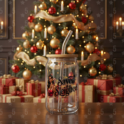Clear glass mason jar with straw featuring 'Saved & Sleighted' design in front of a decorated Christmas tree and presents.