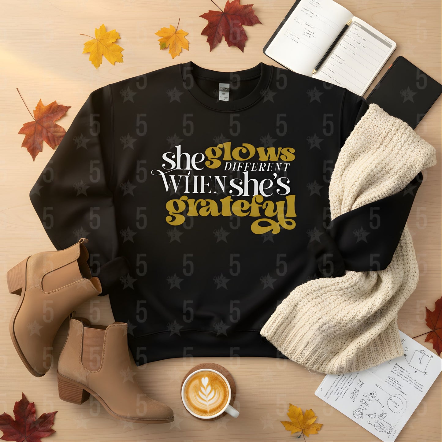 Black sweatshirt with 'she glows different when she's grateful' text, surrounded by autumn leaves, a cup of coffee, and boots on a wooden surface.