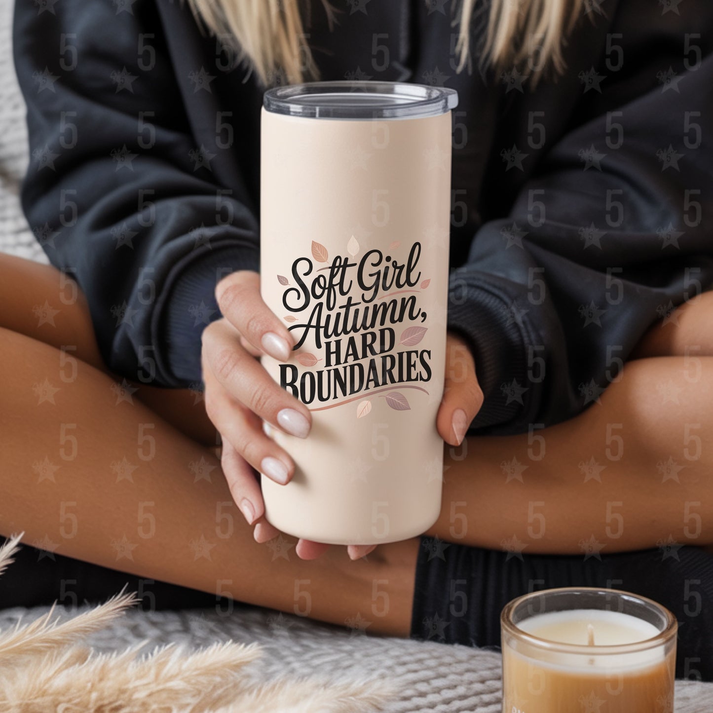 Person holding a tumbler with 'Soft Girl Autumn, Hard Boundaries' text, sitting on a textured surface with a candle.