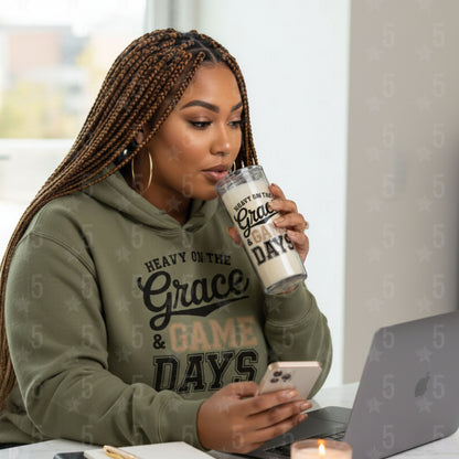 Woman in a green hoodie with text, holding a tumbler and using a phone, sitting at a table with a laptop.
