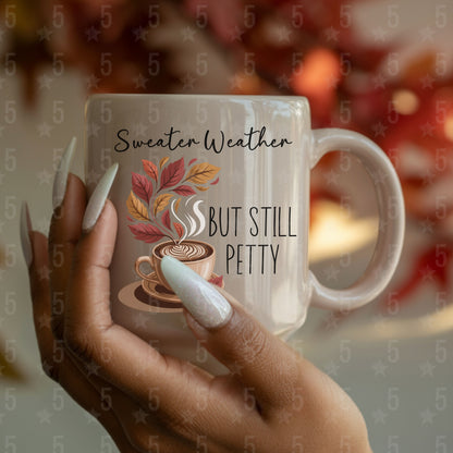 Mug with 'Sweater Weather, But Still Petty' text and autumn design held by a hand.