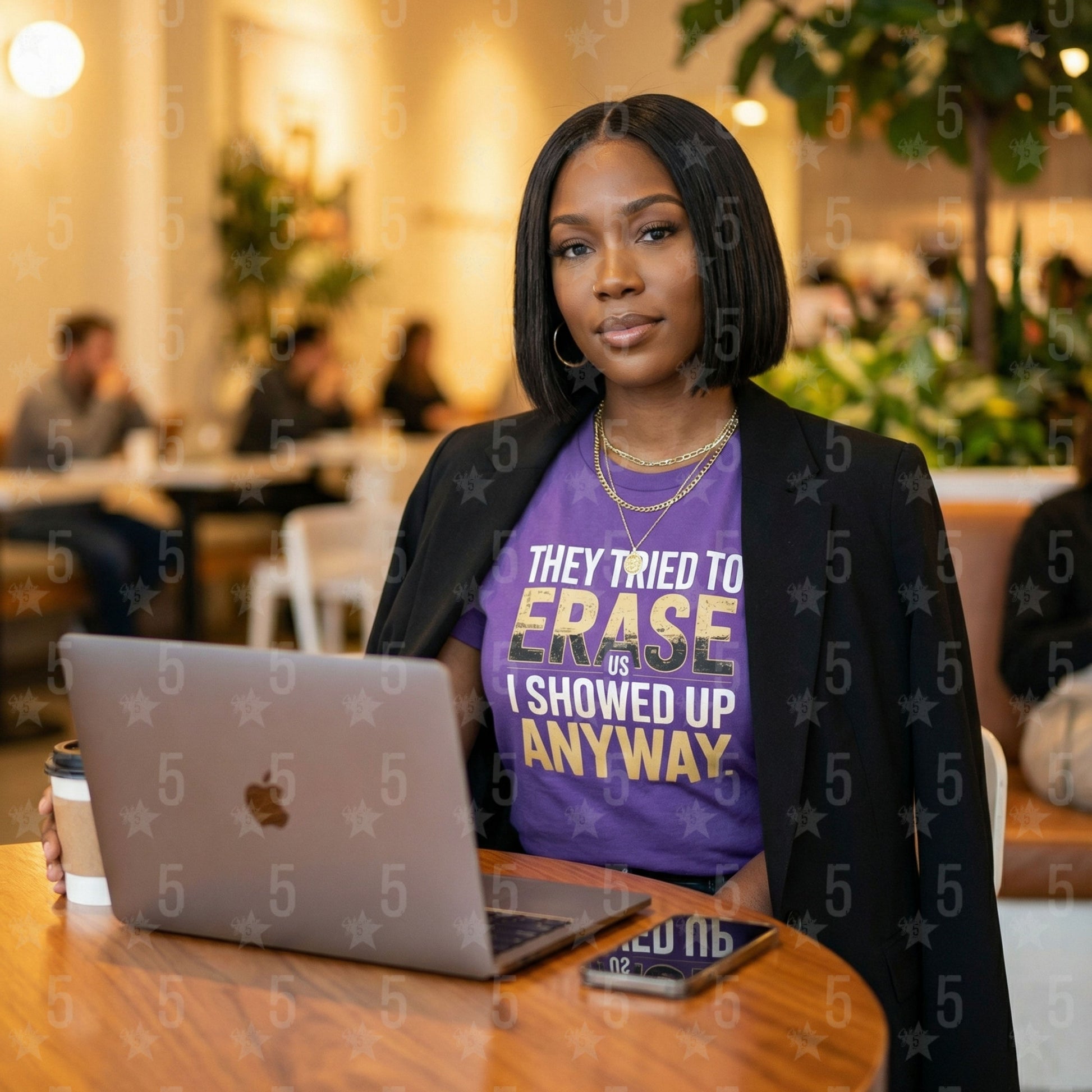 Woman sitting at a table with a laptop, wearing a purple shirt with text, in a casual indoor setting.