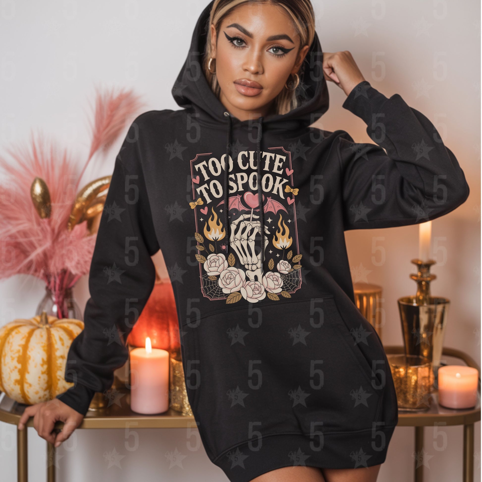 Woman wearing a black hoodie with 'Too Cute To Spook' design in a decorative setting.