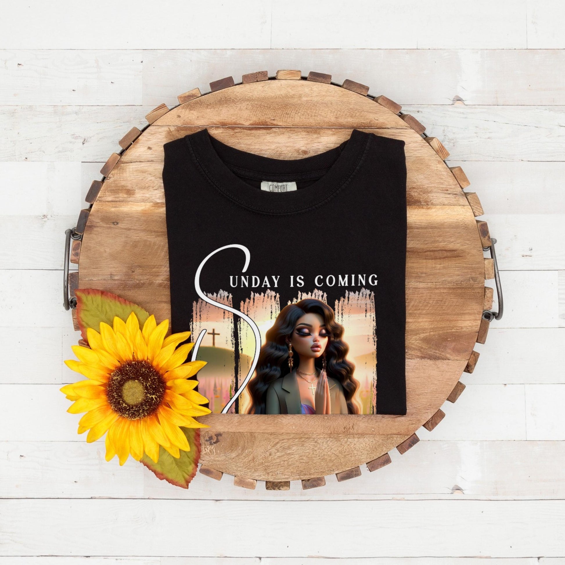 Black folded t-shirt with sunday is coming design on a wooden surface with a sunflower.