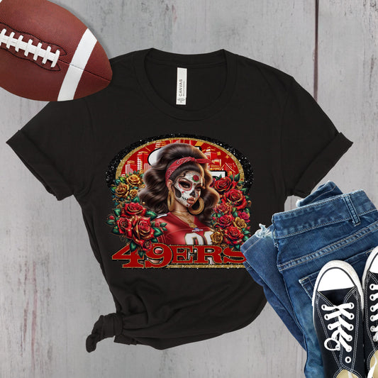 Black t-shirt with a colorful dia de los muertos niners design, football, jeans, and sneakers on a wooden surface.