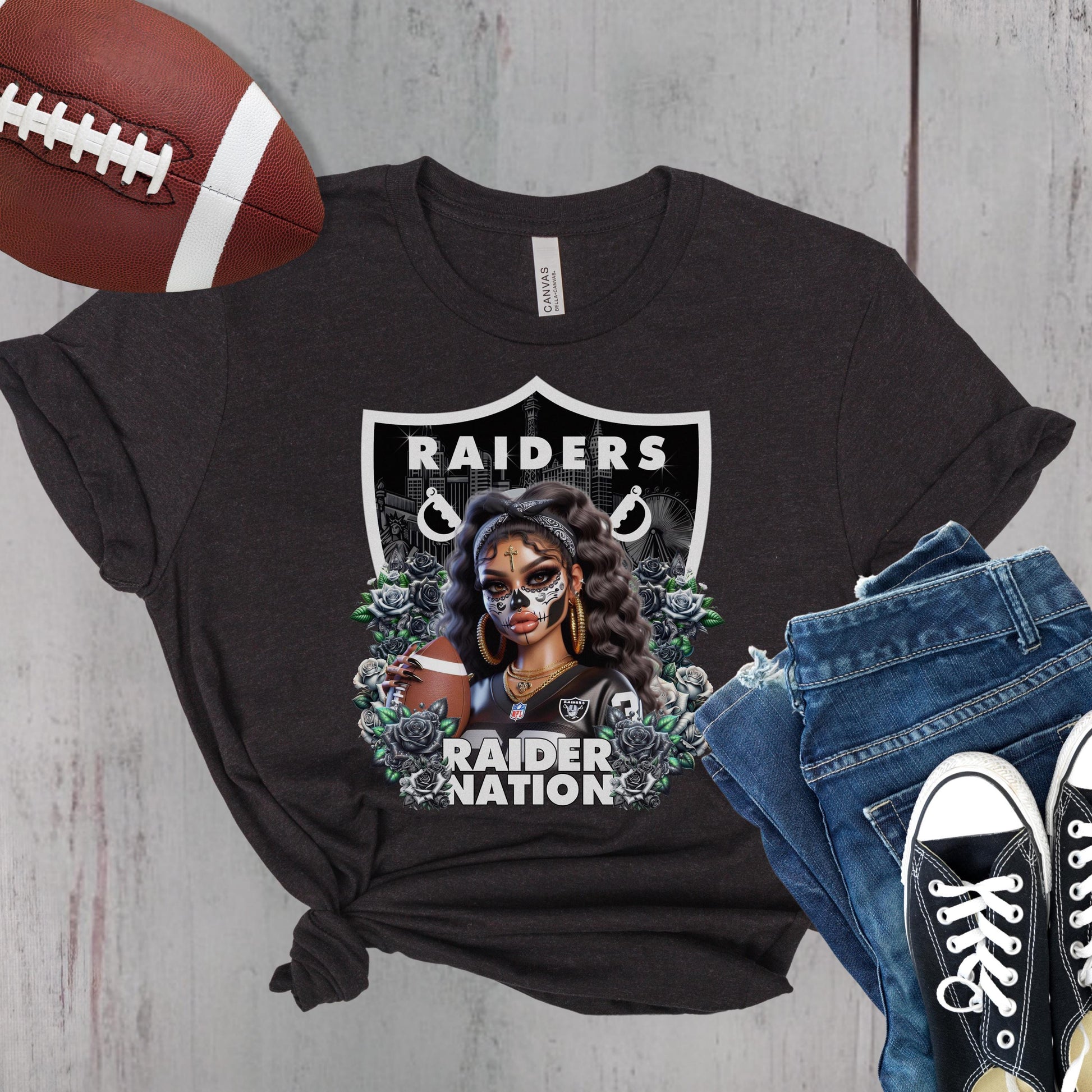 Black t-shirt with a dia de los muertos Raiders design, football, jeans, and sneakers on a wooden surface.