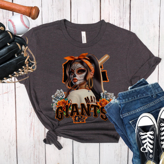 Dark gray t-shirt with a sf giants dia de los muertos design of a woman in a mask and hat, surrounded by roses, on a wooden surface with a baseball, bat, glove, jeans, and black sneakers.