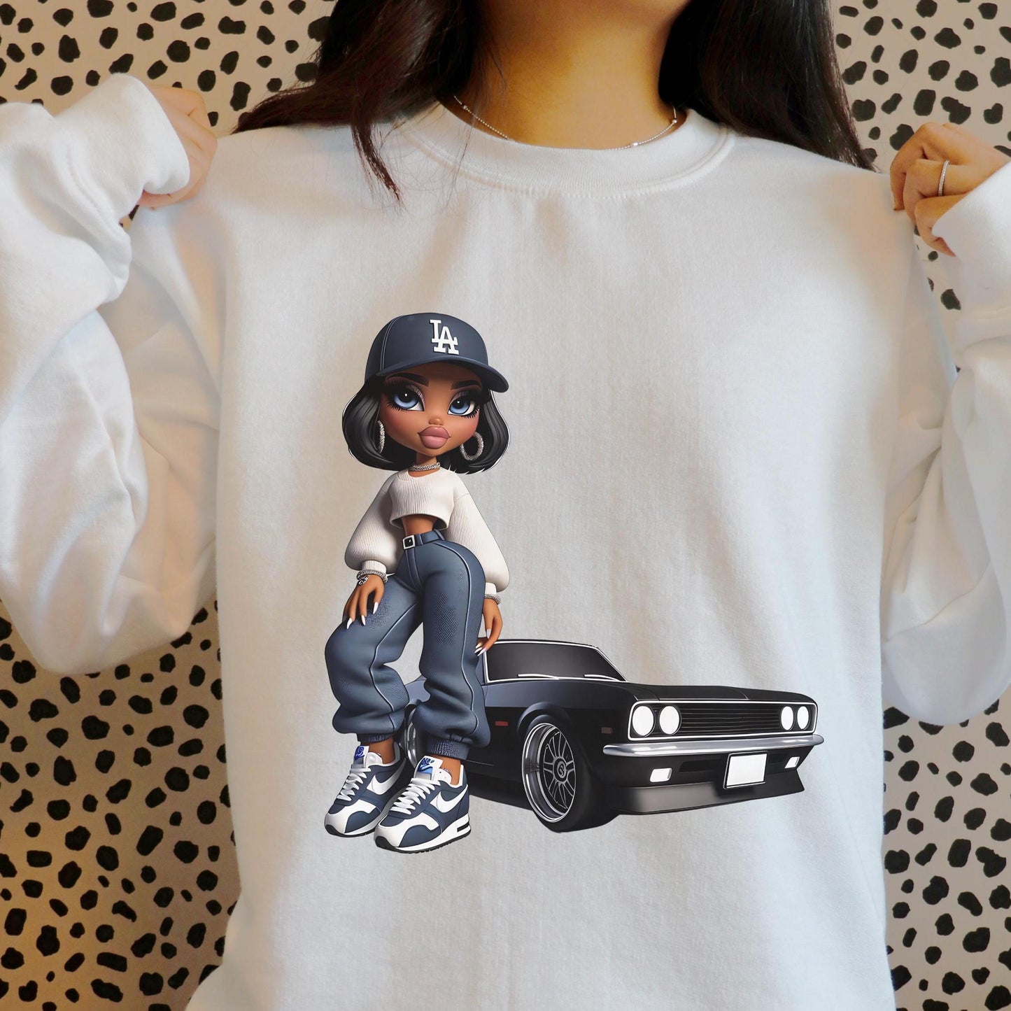 White sweatshirt with a graphic of a bratz doll wearing a cap and jeans, standing next to a car, on a patterned background.