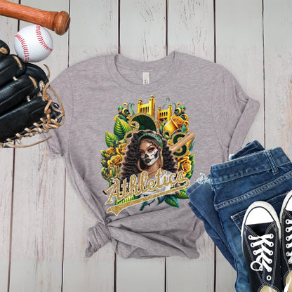 Gray t-shirt with a dia de los muertos athletics design, displayed with a baseball, bat, glove, jeans, and sneakers on a wooden surface.