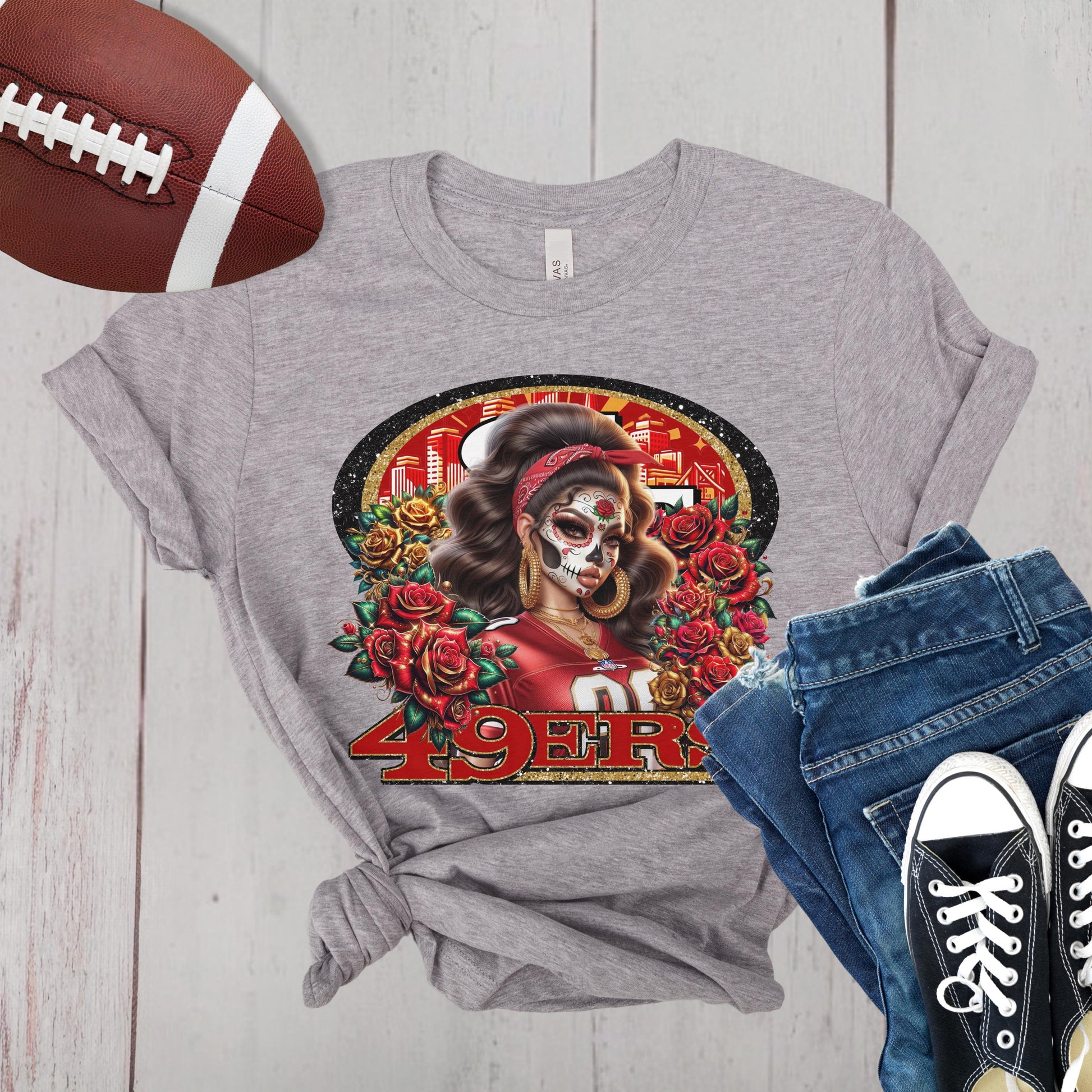 Gray t-shirt with a dia de los muertos niners design, next to a football, jeans, and black sneakers on a wooden surface.