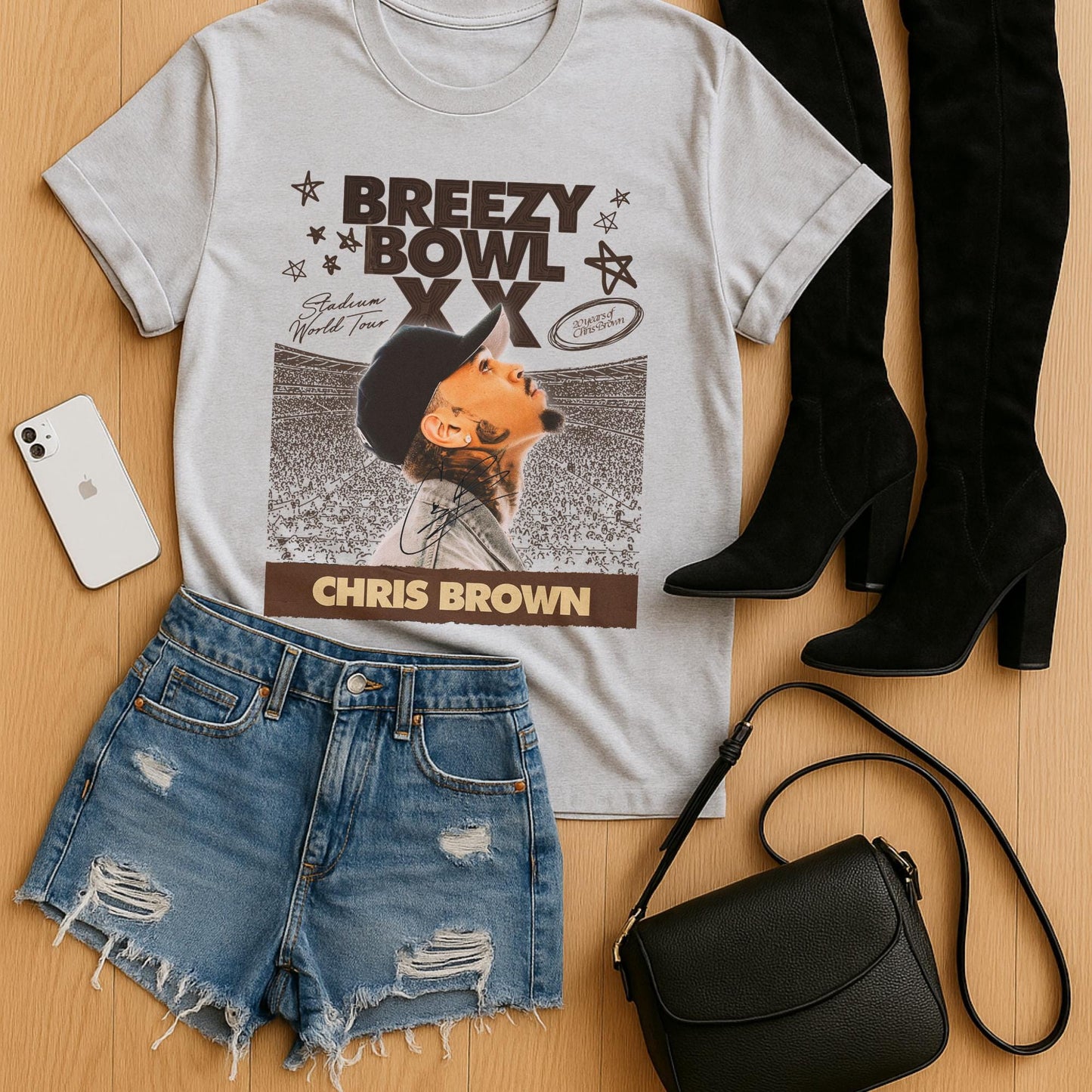 gray T-shirt with Chris Brown graphic, denim shorts, black boots, phone, and black handbag on a wooden surface