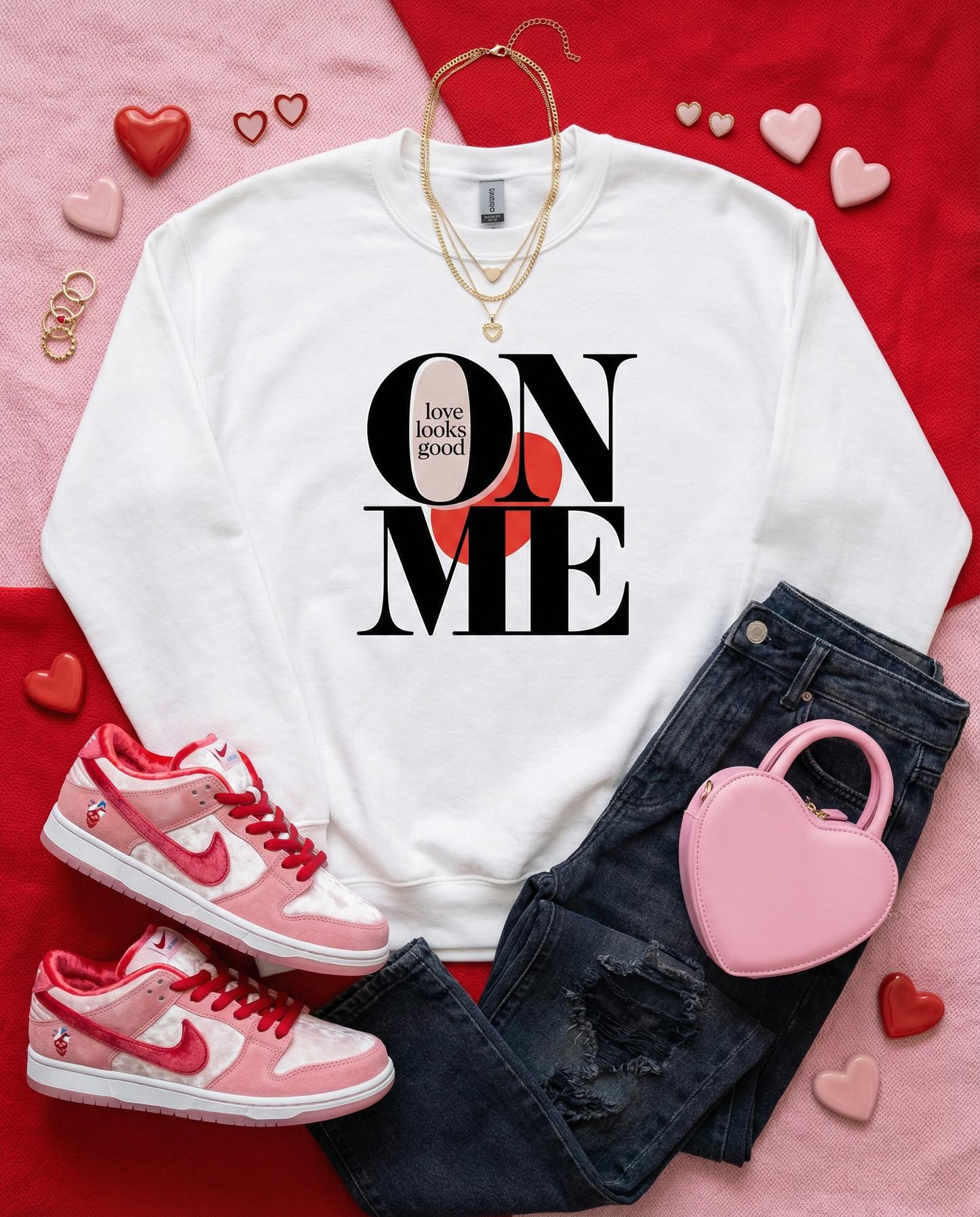 White sweatshirt with 'Love looks good on me' text, pink sneakers, black jeans, and pink heart-shaped handbag on a red and pink background.