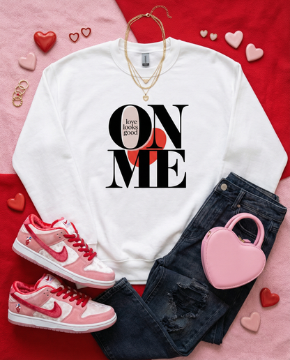White sweatshirt with 'Love looks good on me' text, pink sneakers, black jeans, and pink heart-shaped handbag on a red and pink background.