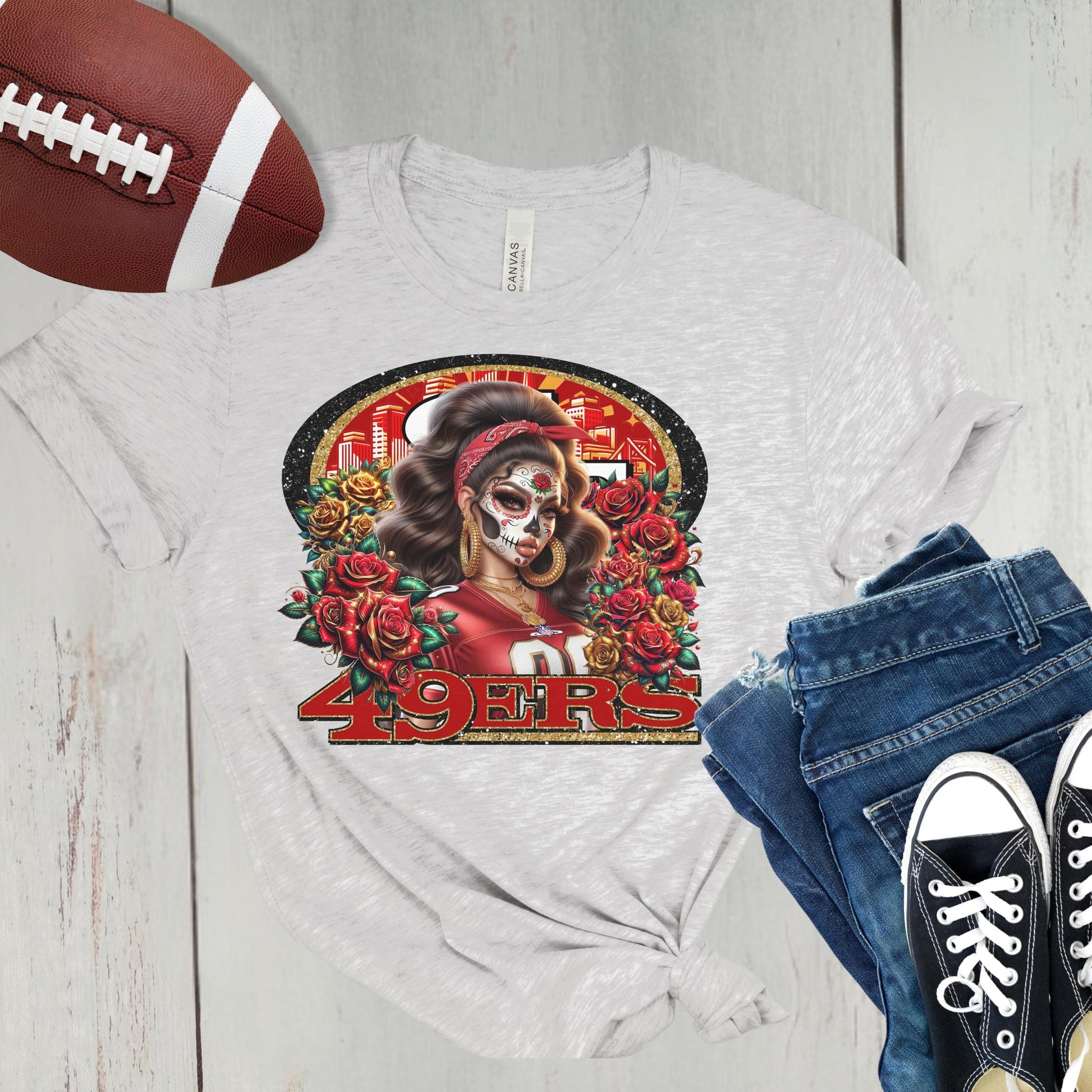 White t-shirt featuring dia de los muertos niners design, next to a football, jeans, and sneakers on a wooden surface.