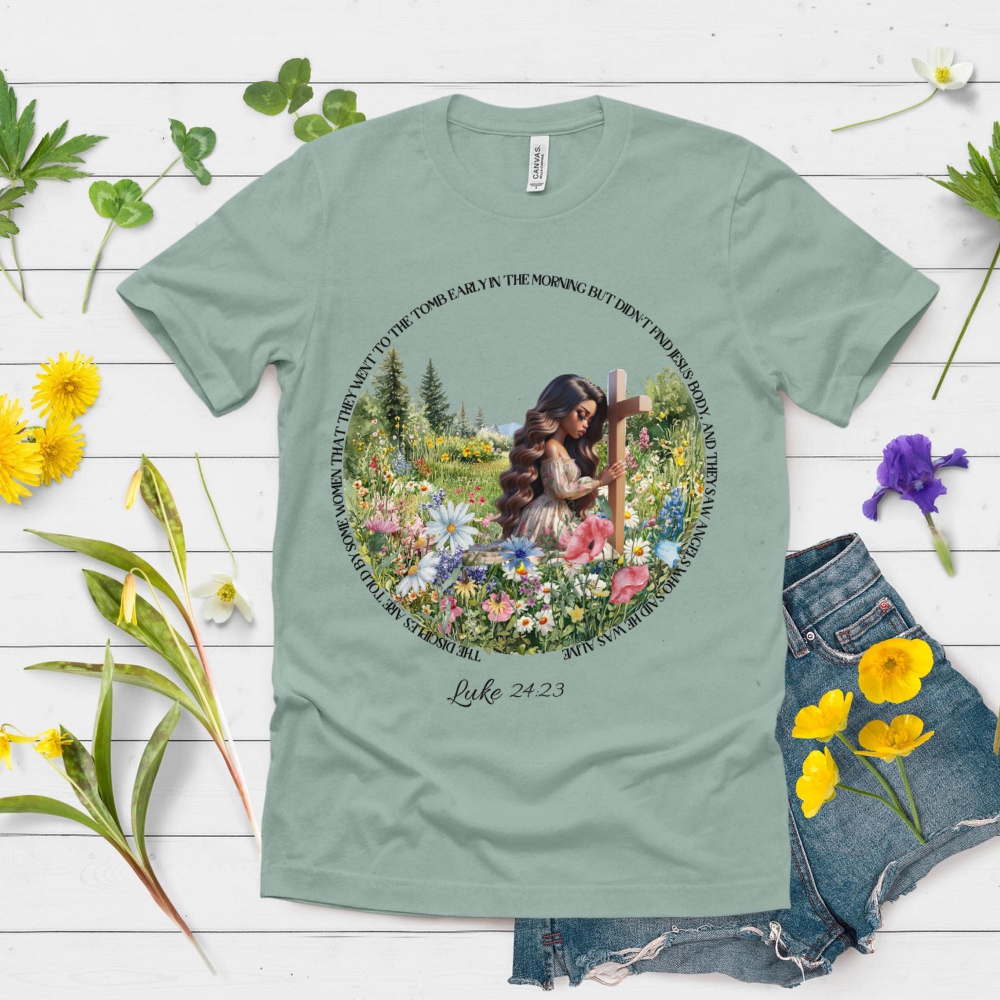 Green t-shirt with a printed design of a woman with flowers and a cross, surrounded by flowers on a wooden surface.